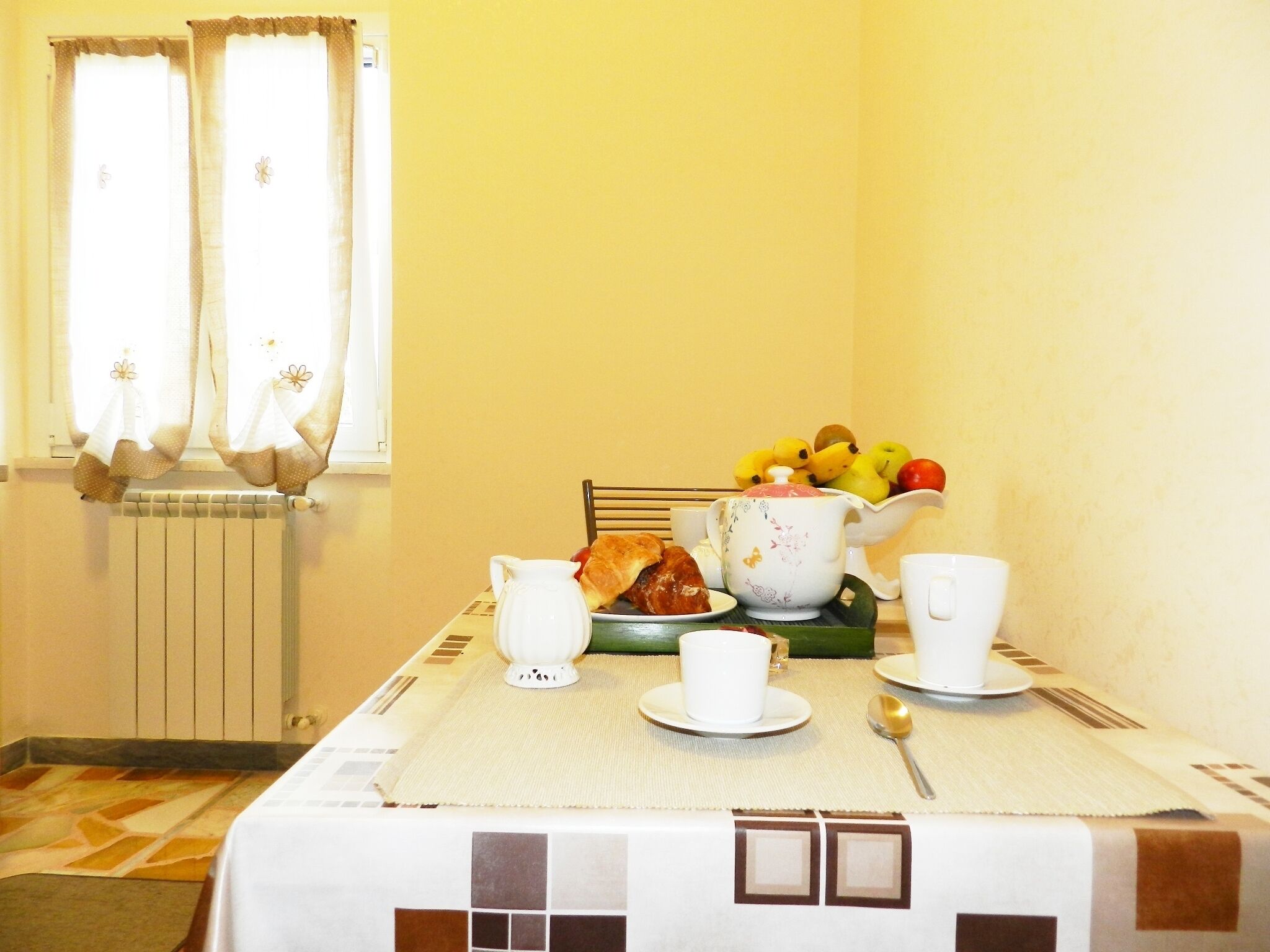 A dining table set with a teapot, cups, a bowl of fruit, and a plate of food.