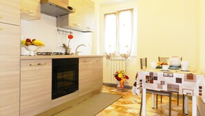 A compact kitchen with a stove, oven, and sink. There's a dining area with a table set for two, a clock on the wall, and a window with curtains. - Anthony's Home 5 people (Roma)