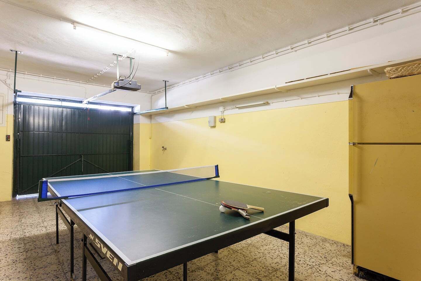 A ping pong table in a room with a closed door, a wall-mounted projector, and a yellow wall.