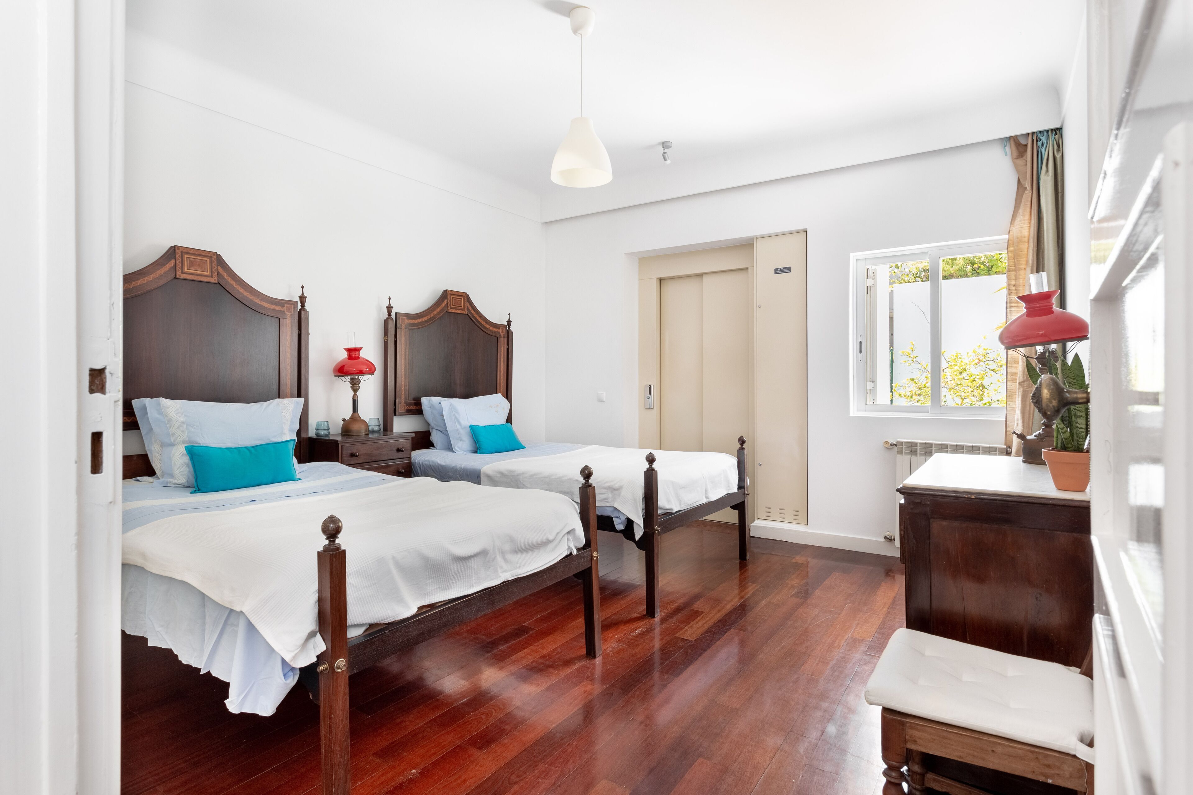 Una habitación con dos camas de madera, ropa de cama blanca, una lámpara roja y una ventana con vistas a la vegetación.