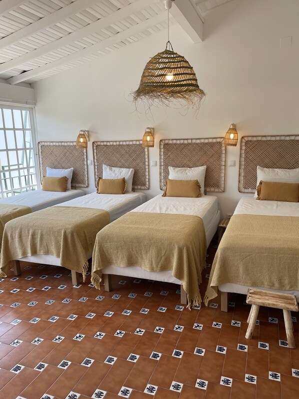 A hotel room with three beds, each with a woven headboard and a beige blanket. There is a woven hanging light fixture above the beds.