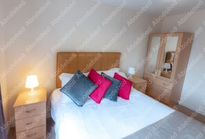 A neatly made bed with pillows, a bedside lamp, a wooden nightstand, and a wardrobe with a mirror.