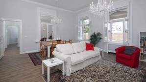 A spacious living room with a white sofa, red armchair, and a chandelier. There is a dining area with wooden chairs and a table, and a bookshelf filled with books. - Grandiose 3BR § 3BathR apt, middle of downtown. Back and better than ever! (Budapest)