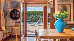 Une pièce avec une table en bois, un vase bleu contenant des plantes vertes, une chaise et une vue sur un espace piscine avec des chaises longues et des parasols.