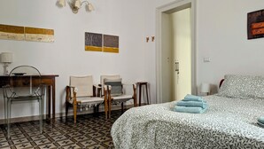 Una habitación de hotel con cama, dos sillas, un escritorio con lámpara y una pared decorada con obras de arte.