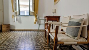 Una habitación con piso estampado, una silla de madera, una mesita y una ventana con cortinas.