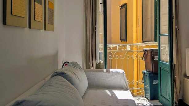 Una habitación estrecha con cama, ventana con cortinas y vistas a un balcón.