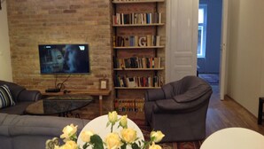 Una sala de estar con chimenea de ladrillo, un televisor de pantalla plana, una estantería, dos sofás grises y una mesa con un jarrón de rosas amarillas.