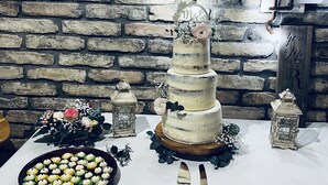 Un pastel de bodas de varios niveles con un adorno decorativo, rodeado de velas y flores sobre una mesa con un fondo de pared de piedra.