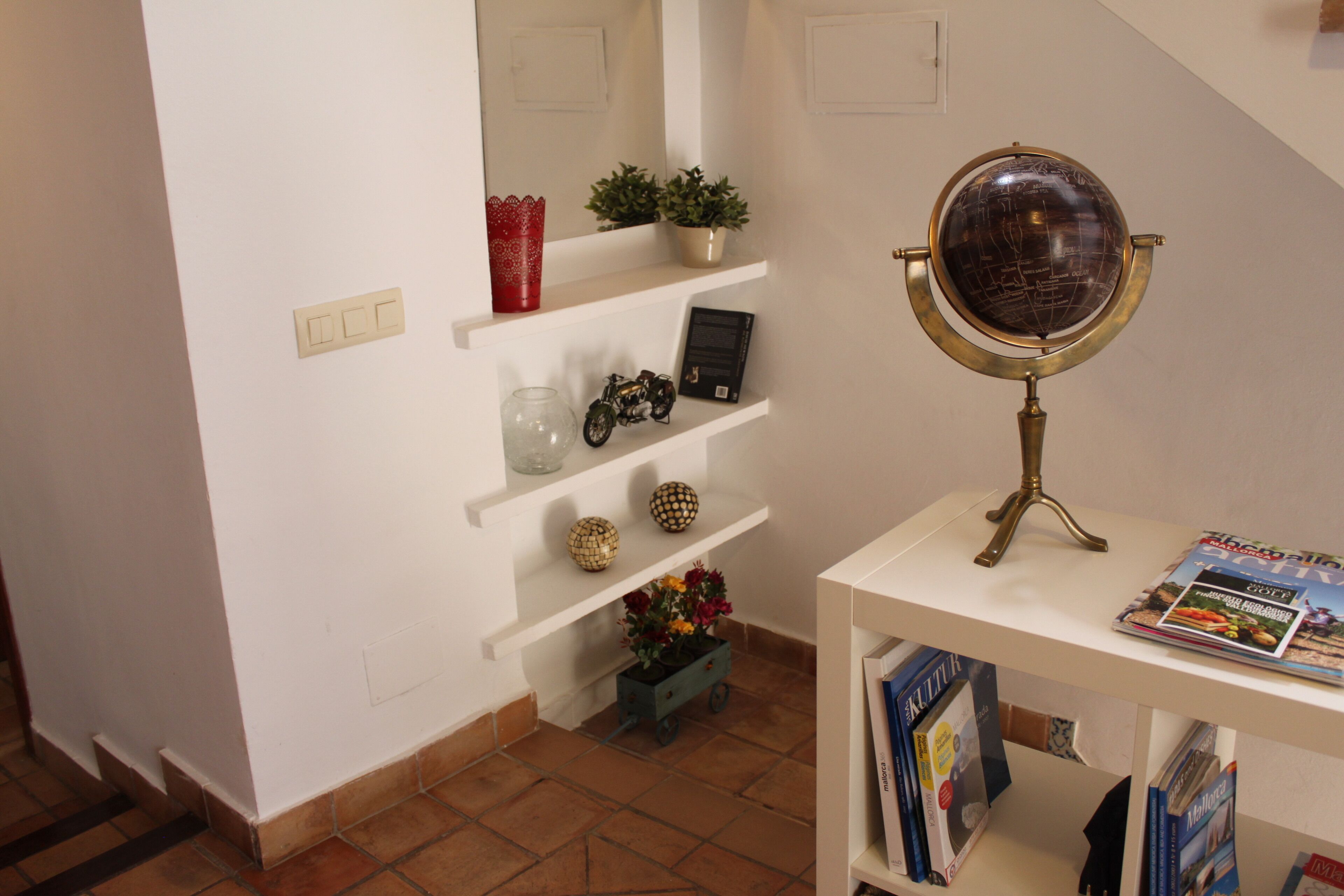 Une pièce avec un mur blanc, une étagère ornée d’objets décoratifs, un globe terrestre posé sur un socle et un petit chariot rempli de fleurs.