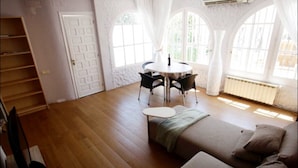 Una habitación amplia con suelo de madera, una mesa redonda con sillas, un sofá y ventanas arqueadas grandes.