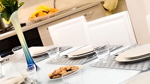 A dining table set with plates, a clear glass holder with cookies, and a blue object, possibly a water bottle, in a modern kitchen with white cabinets and a yellow flower in the background. - GREGORY BIG FAMILY (Roma)