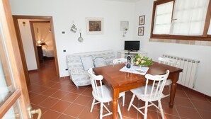 Uma área de refeições com uma mesa de madeira e cadeiras brancas, um sofá com almofadas estampadas, uma televisão e um radiador.