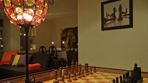 Un jeu d’échecs est installé sur une table en bois dans une pièce ornée d’un lustre, d’un tableau au mur et d’un canapé avec des coussins.