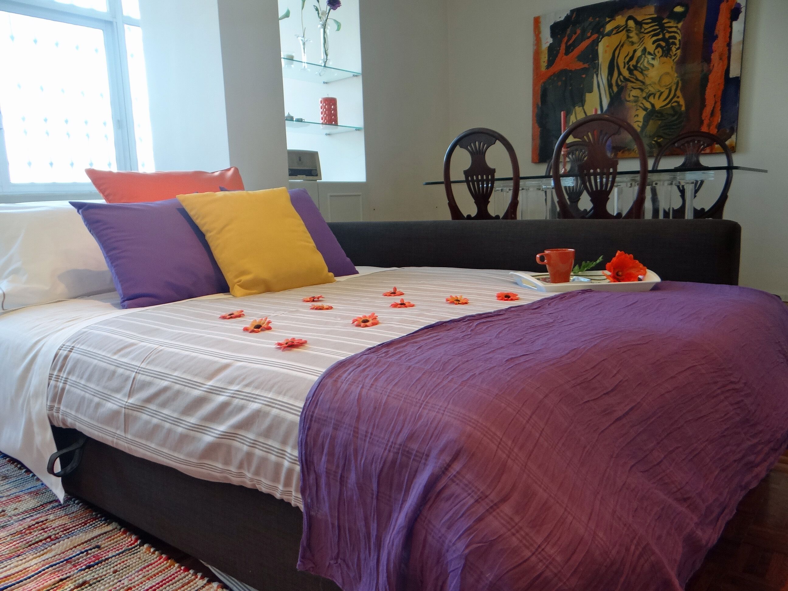 Cama bem arrumada com colcha listrada, manta roxa e flores laranjas. Há duas cadeiras de madeira e um quadro de uma tigresa na parede.