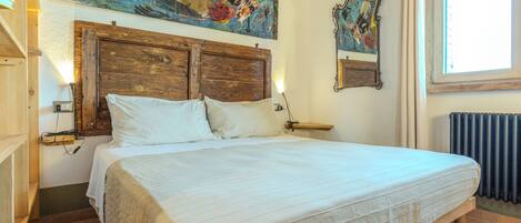 A bedroom with a wooden headboard, a large bed with white linens, a painting above the bed, a mirror on the wall, and a window with curtains.