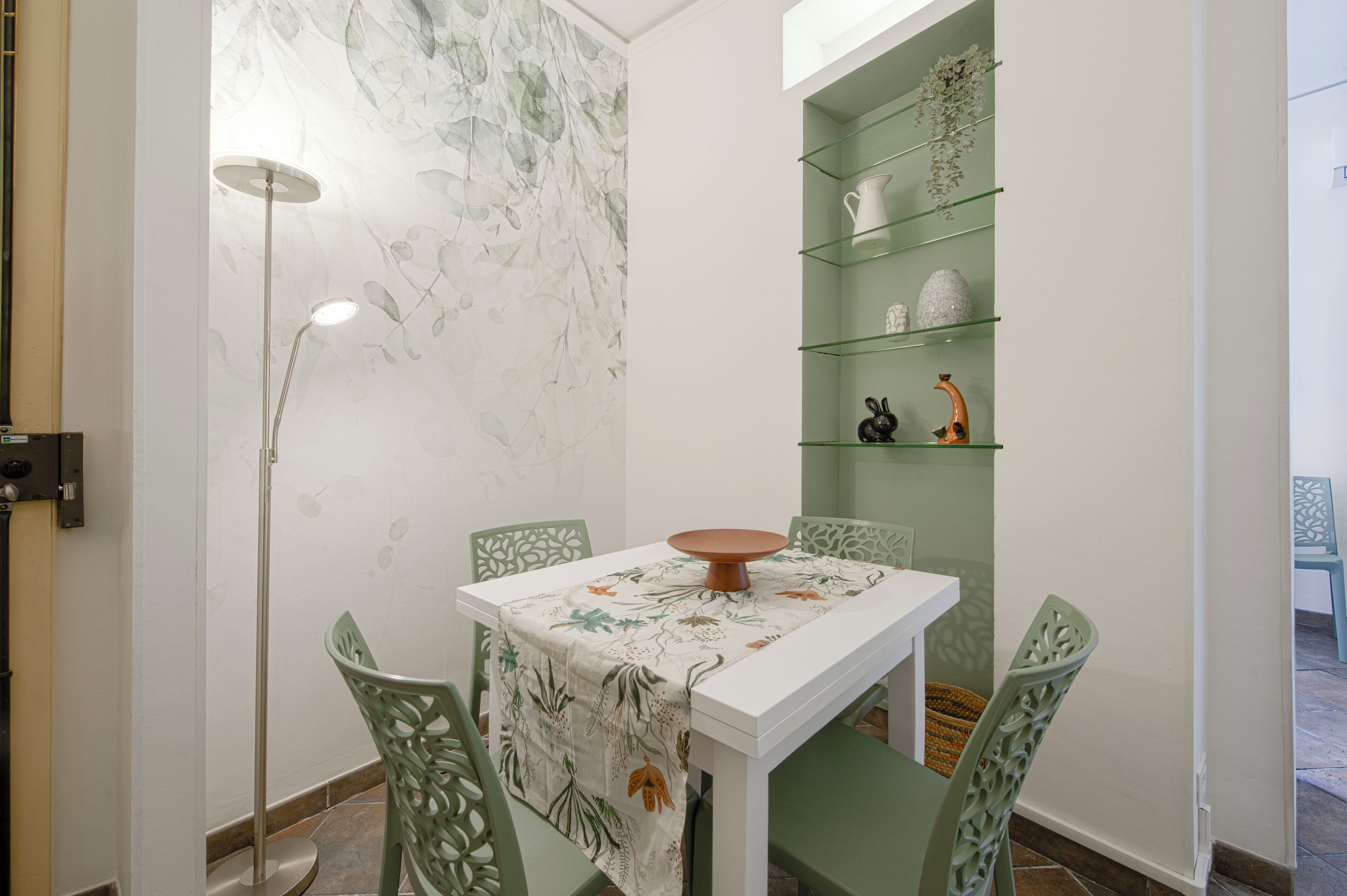 A dining area with a white table, green chairs, a floor lamp, and a green shelving unit with decorative items.
