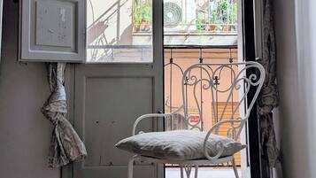 Ein Zimmer mit einem gemusterten Boden, einem weißen Stuhl und einer offenen Tür, die auf einen Balkon mit Blick auf ein Gebäude und Grünflächen führt.