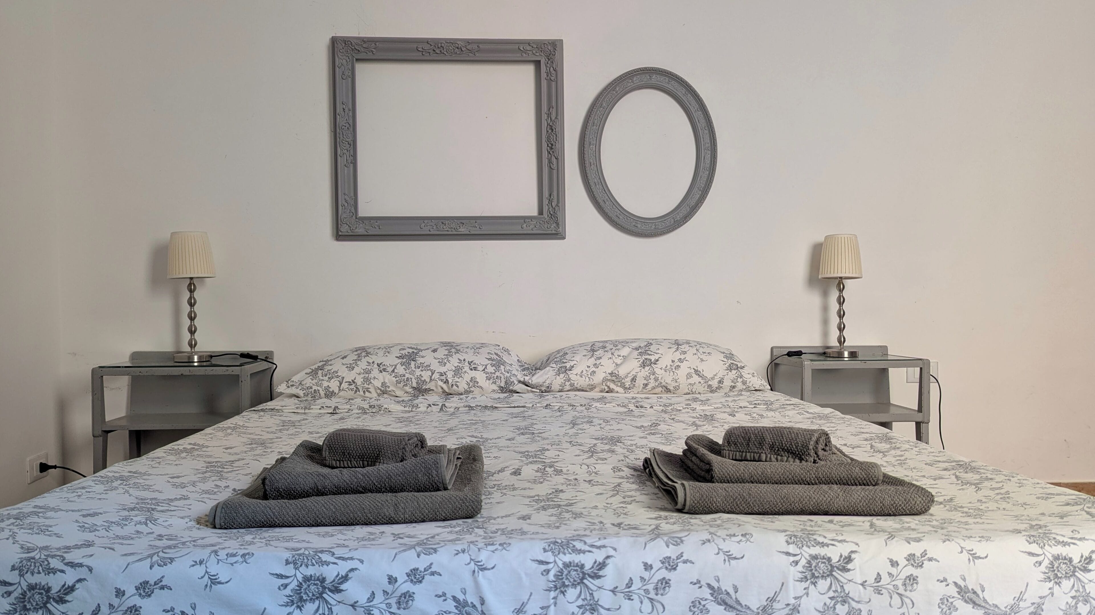 Ein ordentlich bezogenes Bett mit floralem Bettbezug, zwei Nachttische mit Lampen und ein dekorativer Spiegel an der Wand.