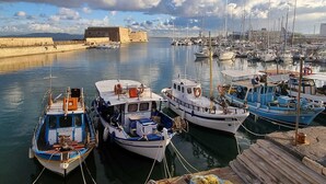 Una marina con diverse imbarcazioni ormeggiate, un forte in pietra sullo sfondo e cielo sereno.