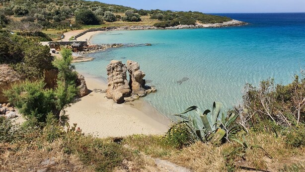Paesaggio costiero con acque cristalline turchesi, formazioni rocciose e una spiaggia sabbiosa.