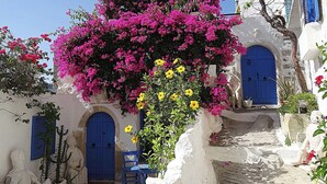 Un edificio bianco con porte e finestre blu, una sedia azzurra e fiori di bouganvillea rosa.