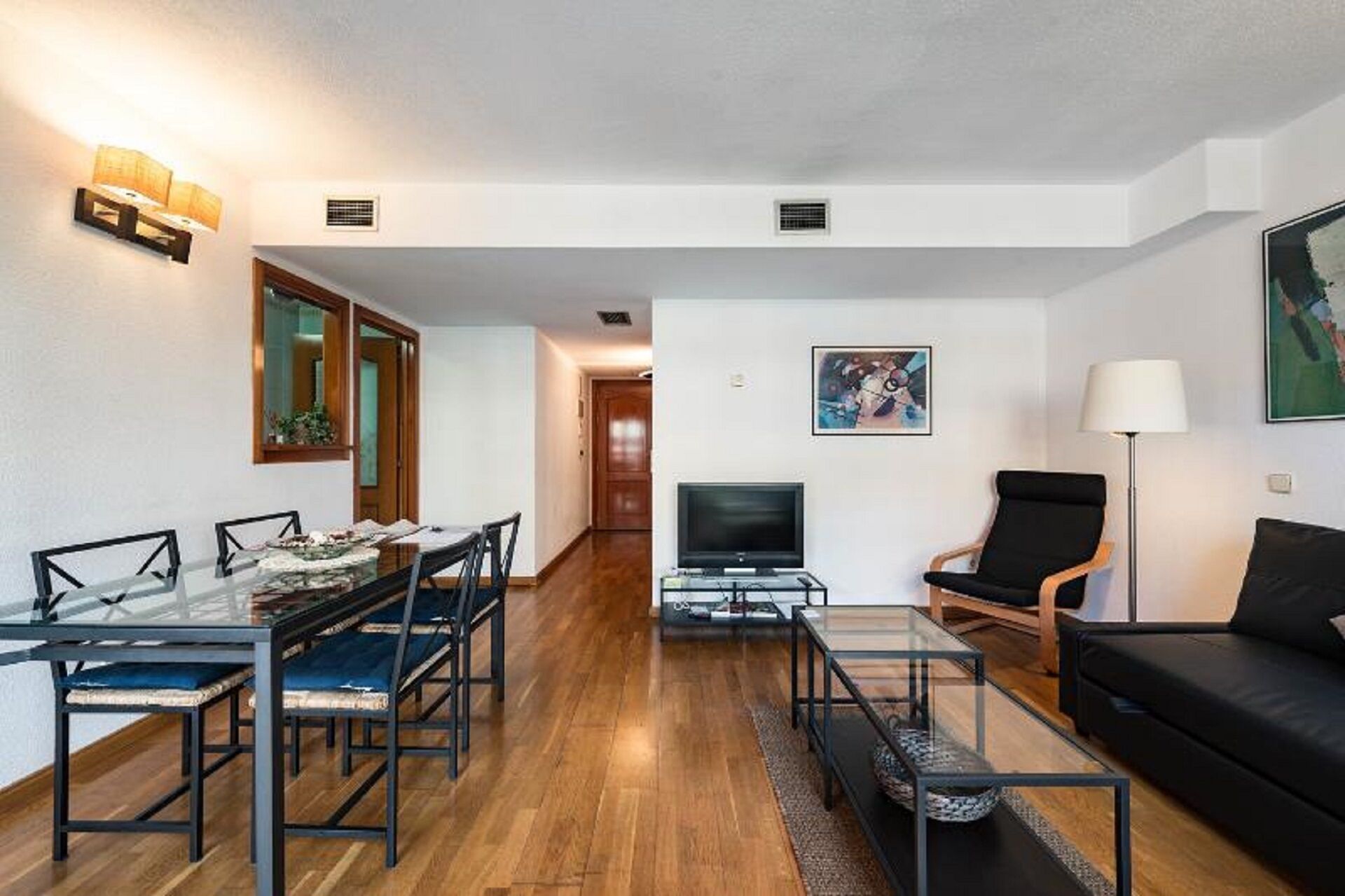 A modern living room with a glass dining table, black sofas, and a flat-screen TV.