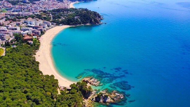 Vista aérea de una zona costera con playa de arena, aguas azules cristalinas y una zona urbana densamente construida al fondo.