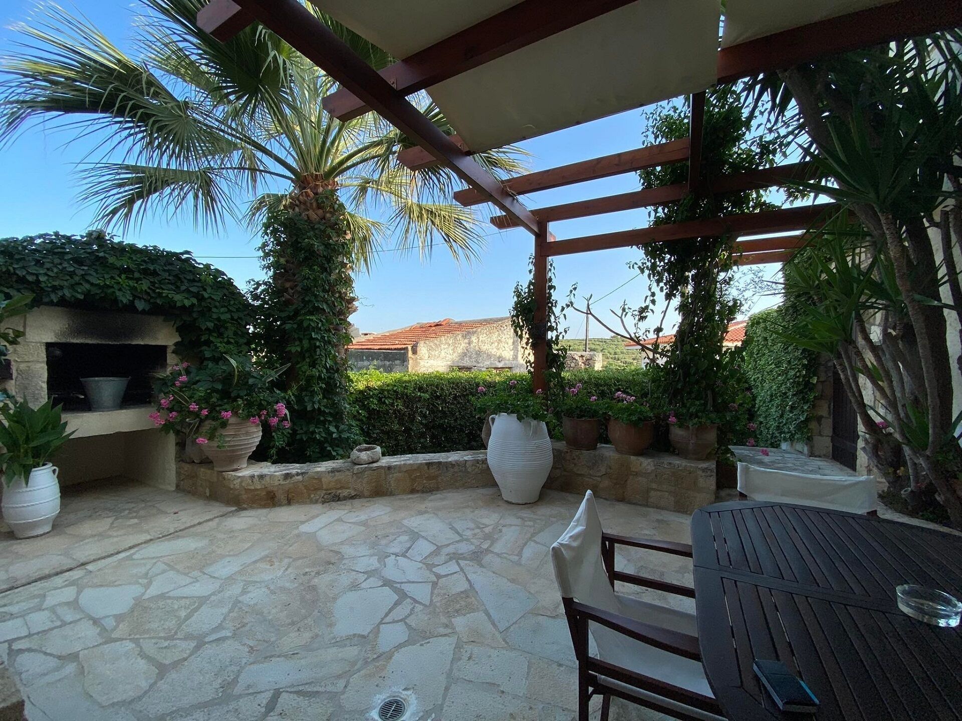 A stone patio with a pergola, outdoor seating, a grill, and potted plants.