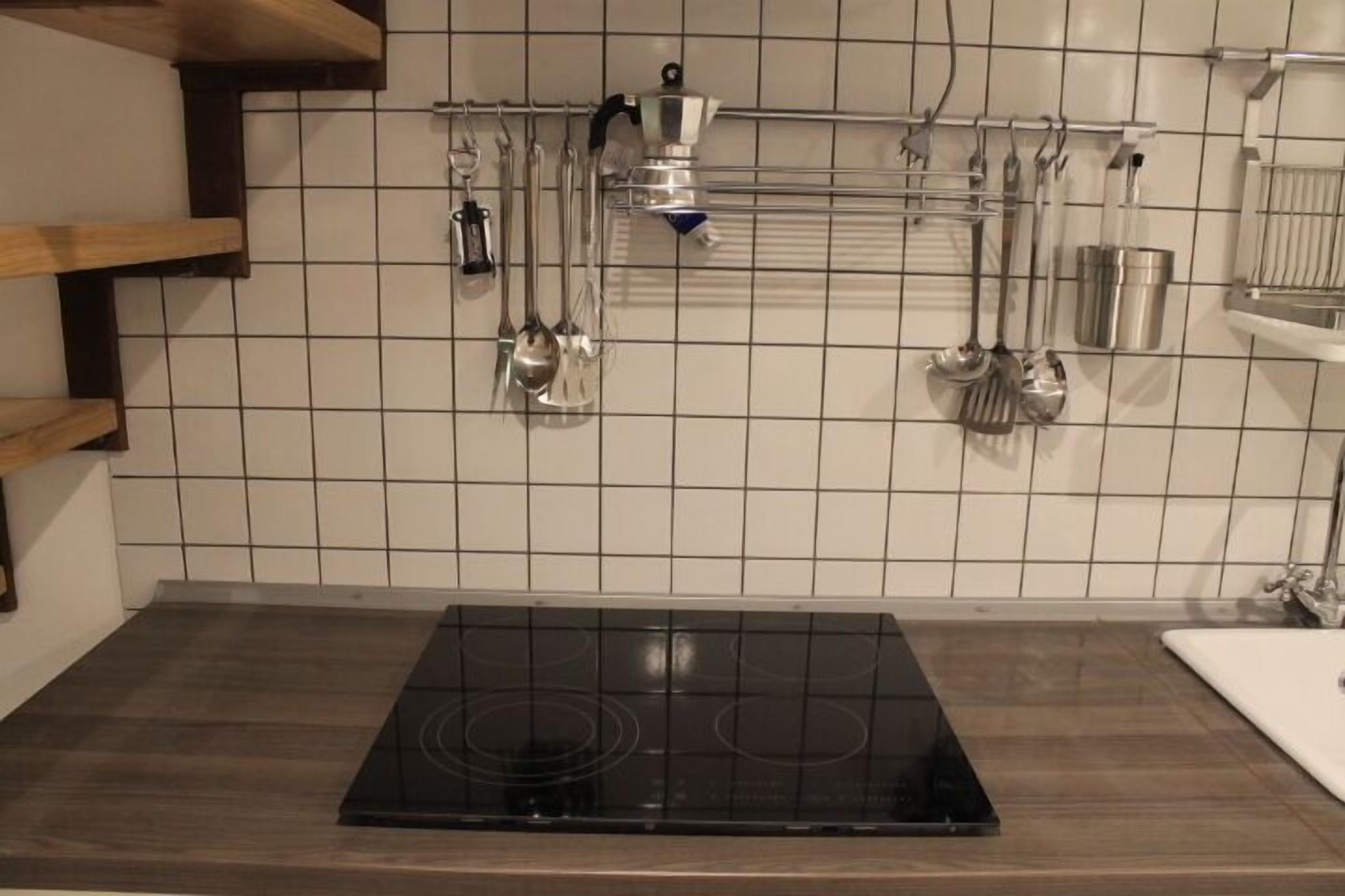 Eine Küche mit einem schwarzen Induktionskochfeld, einer weißen Fliesenrückwand und verschiedenen Küchenutensilien, die an einem Gestell hängen.
