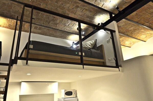 Un dormitorio tipo loft con cama, una kitchenette pequeña y vistas a la ciudad a través de un ventanal amplio.