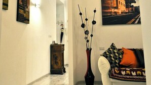 A hallway with a white wall, a wooden floor, a black metal beam ceiling, a white sofa with a patterned throw, a vase with branches, and a painting of a building. - Quiet luxury apartment in the middle of Rome historical center. (Roma)