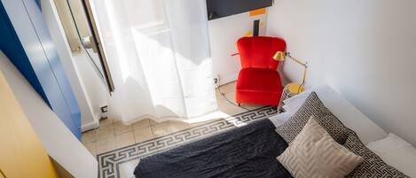 Ein Zimmer mit einem Bett, einem roten Sessel, einem Schreibtisch mit Lampe und einem großen Fenster mit Vorhängen.