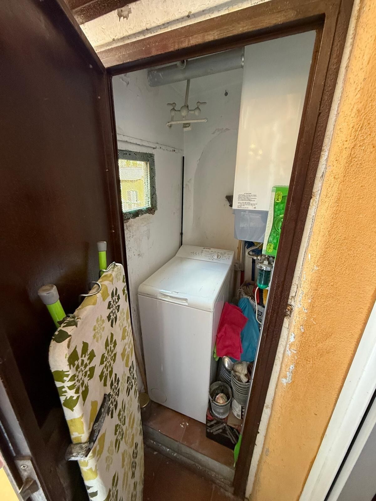 A compact laundry room with a washing machine, a small window, and various cleaning supplies.