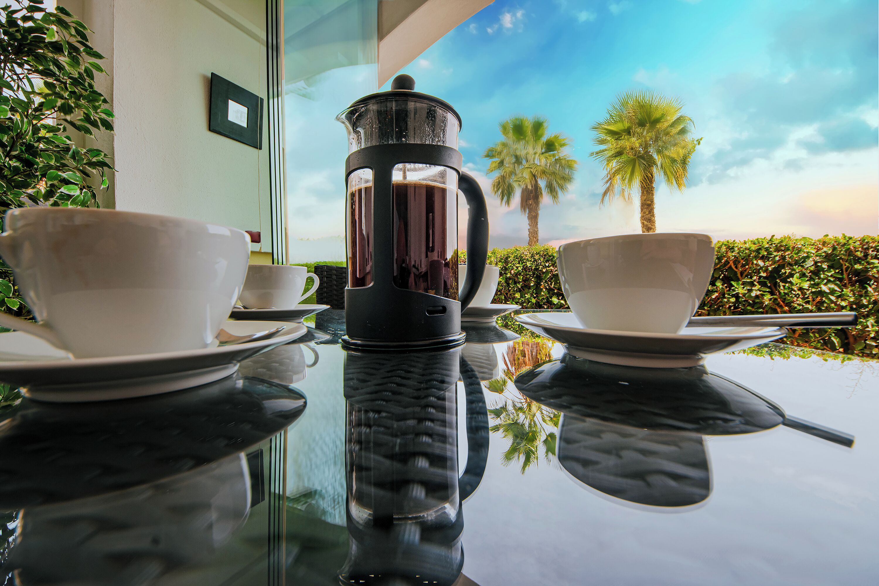 Un tavolo con due tazze bianche e una macchina da caffè nera, con il riflesso di una palma e del cielo sullo sfondo.