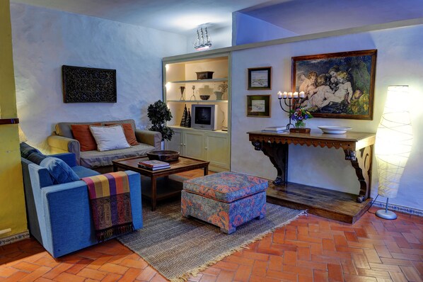 A living room with a sofa, ottoman, coffee table, and a television. There is a painting on the wall and a lamp on a side table.