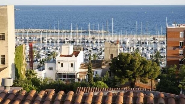 A marina with numerous boats docked, a coastal building with a tiled roof, and a view of the sea.