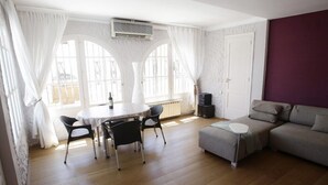 A room with a round table and chairs, a sofa, a radiator, and a window with curtains.