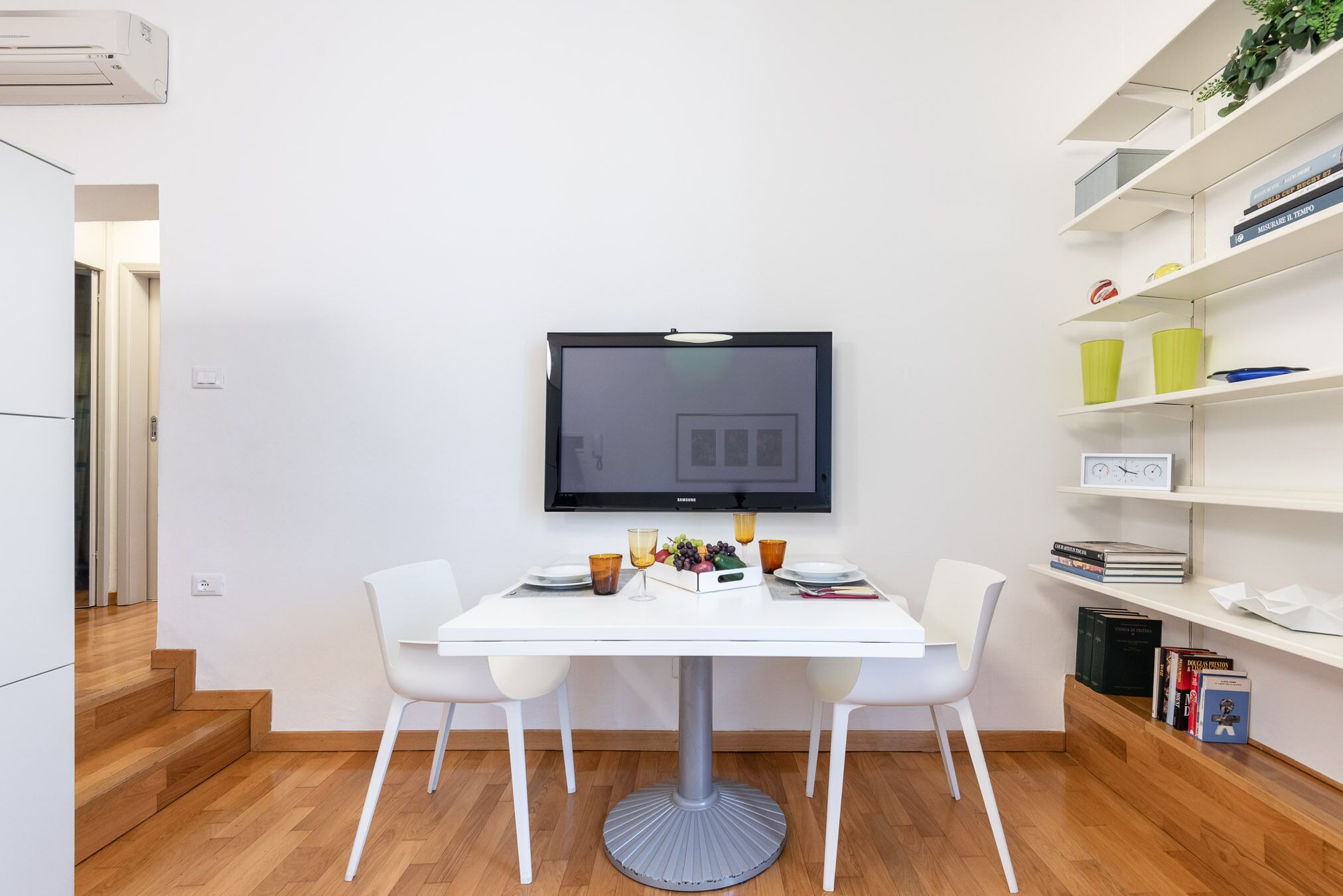 Un espace repas moderne avec une table blanche, deux chaises blanches et un téléviseur fixé au mur.