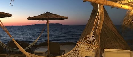 A beach scene with a hammock, chairs, and a thatched roof structure.
