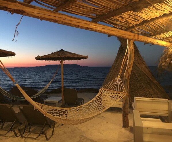 A beach scene with a hammock, chairs, and a thatched roof structure.
