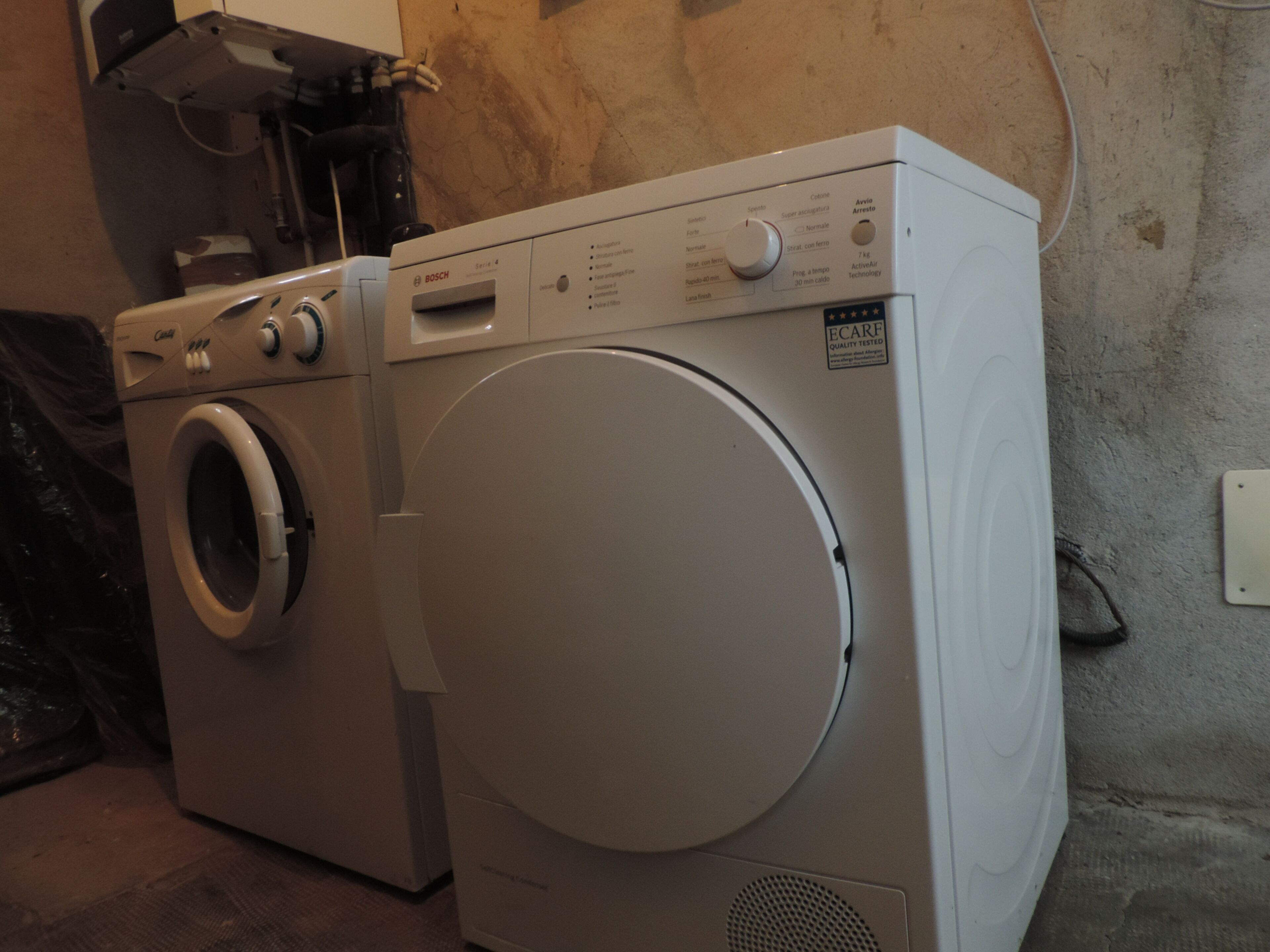 A front-loading washing machine with a control panel, a dryer, and a wall-mounted electrical outlet.