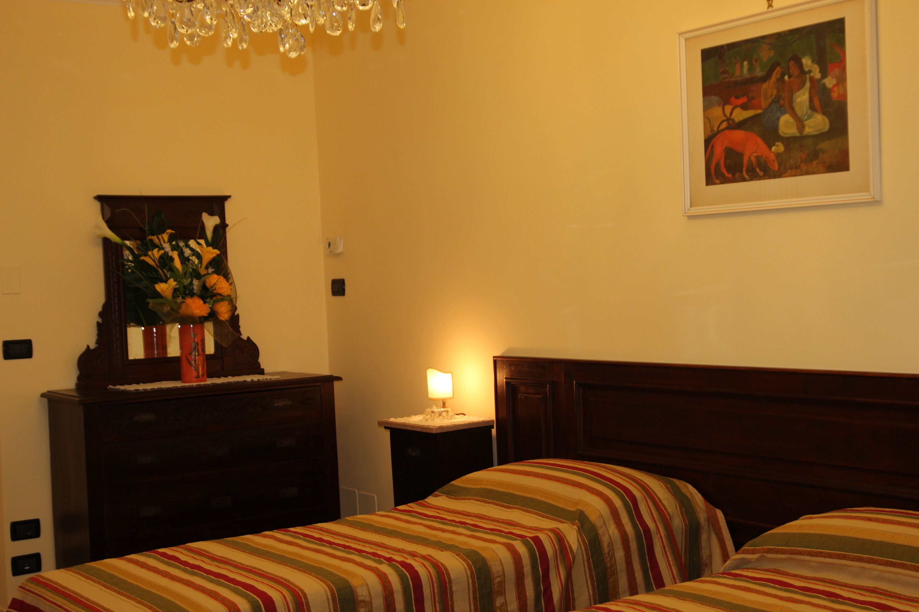 A hotel room with a bed, a dresser, a nightstand with a lamp, a painting on the wall, and a chandelier.