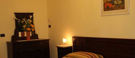 A hotel room with a bed, a dresser, a nightstand with a lamp, a painting on the wall, and a chandelier.