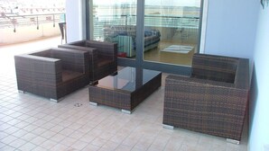 A balcony with wicker furniture, a glass table, and a view of the sea and buildings.