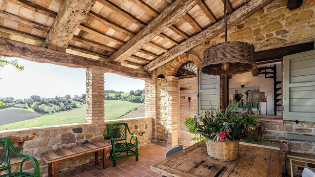 Un patio rústico de piedra con una mesa de madera, sillas verdes y una cesta de mimbre con flores.