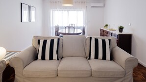 Sala de estar com sofá bege, mesa de jantar com cadeiras, área de cozinha com armários e uma grande janela com cortinas leves.