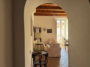 A hallway with a wooden ceiling, a lamp, a table with a basket and plant, and a chair with a floral pattern.