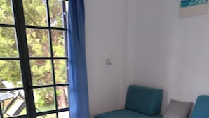 A room with a white tiled floor, a blue sofa with cushions, a white side table with papers and a small plant, and a window with a blue curtain. - Julie's studio in Kokkini Hani, 2 mins walk to beach. Near famous theme parks!  (Kokkini Hani)