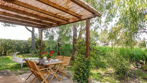 Uma pérgola de madeira com uma mesa e cadeiras, cercada por vegetação e um arbusto.